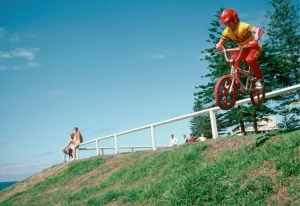 Un jeune biker franchit une barrière au bord de mer avec son BMX rouge, dans le film BMX Bandits.