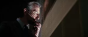 Le méchant Alan Rickman pensif, devant une fenêtre de nuit ; une lueur blanche sort à travers les minces espaces du store fermé devant lui ; plan issu du film Die Hard : Piège de Cristal.