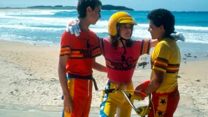 Nicole Kidman entre ses deux camarades de bikers, chacun de ses bras sur les épaules des deux garçons ; ils sont en tenue de cyclistes et sont sur la plage, on voit l'océan derrière, et le sable ; plan issu du film BMX Bandits.