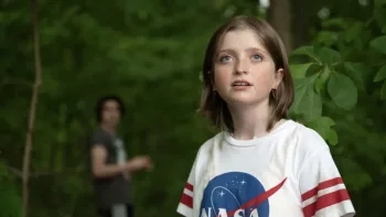 En forêt, une jeune fille avec un t-shirt Nasa observe le ciel avec une attitude de surprise lumineuse ; en flou derrière elle, un jeune homme ; plan issu du film Le monde est à nous.