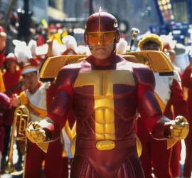 Arnold Schwarzenegger en costume de super-héros flashy rouge et or prêt à intervenir en tête d'une grande fanfare à ses couleurs, qui l'acclame, dans le film La course aux jouets.