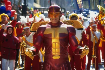 Arnold Schwarzenegger en costume de super-héros flashy rouge et or prêt à intervenir en tête d'une grande fanfare à ses couleurs, qui l'acclame, dans le film La course aux jouets.
