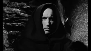 Plan rapproché-épaule sur un homme en capuche, devant une paroi de pierre, l'air sombre, dans le film Horror Hotel.