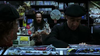 Au premier plan un prêtre fouille dans les bacs d'un magasin de vinyle ; derrière lui, un homme chevelu le fixe avec un air sombre ; plan issu du film Le jour de la bête.