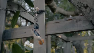 Un porte-clé représentant une chaussure Converse pend à une barrière en bois dans un jardin ; au sommet du porte-clé, on distingue la toute petite silhouette de Marcel le coquillage.