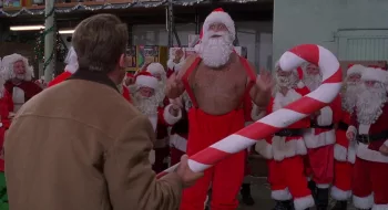 Arnold Schwarzenegger, vu de dos, s'apprête à frapper tout un groupe de faux Pères Noël avec un sucre d'orge géant dans le film La course aux jouets.