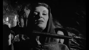 Une femme d'âge mûr, encapuchonnée, tient un couteau sous sa gorge, l’air menaçant, dans ce qui semble être une grotte, seulement éclairée par des bougies ; plan issu du film Horror Hotel.