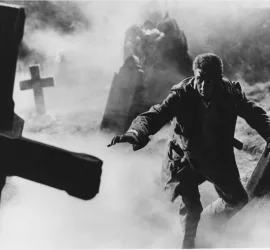 Un homme avance à tâtons dans un cimetière aux croix de travers et plongé dans une brume épaisse ; plan du film Horror Hotel.