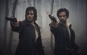Romain Duris et François Civil dégainent leur pistolet en pleine forêt, plongée dans la grisaille ; scène du film Les Trois Mousquetaires : Milady.