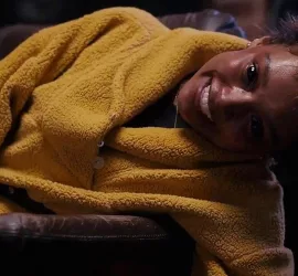 Une jeune femme est effondrée sur un canapé dans un peignoir jaune, le visage figé dans un rictus inquiétant ; scène du film La Main produit par A24.