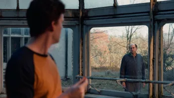 Au premier plan, un jeune homme de profil la tête tournée vers Nicolas Cage, au second plan, qui l'observe les bras ballants, derrière une vitre qui donne sur de la verdure ; plan issu du film Dream Scenario.