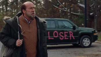 Nicolas Cage, sac en bandoulière aborde une forêt l'air soucieux ; derrière lui est garée une voiture verte sur laquelle on a taggé en rose le mot "Loser" ; plan du film Dream Scenario.