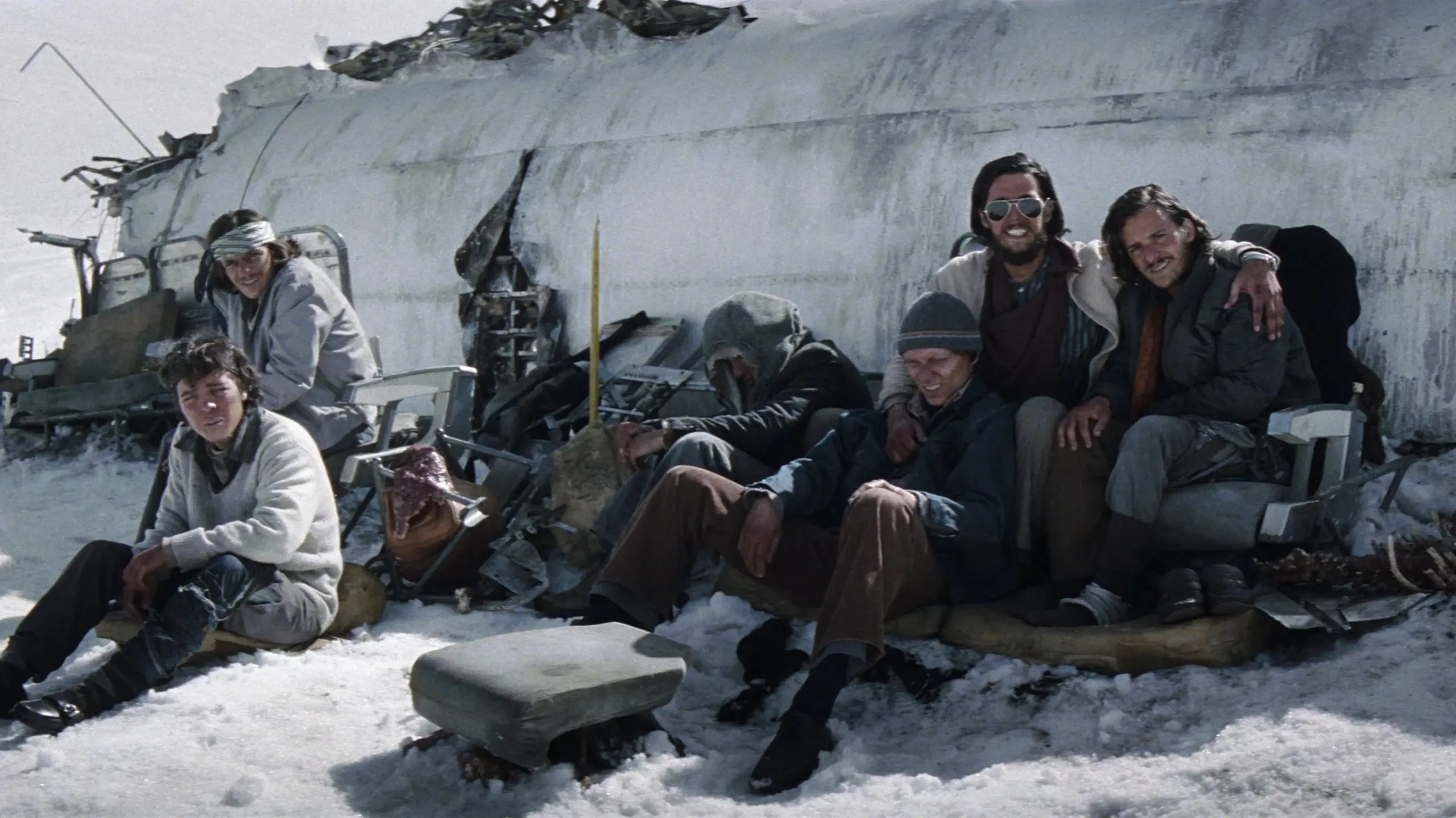 Tout le groupe de survivants d'un crash aérien dans les Andes du film Le cercle des neiges, posent assis contre l'épave de leur avion, les uns à côté des autres, certains avec le sourire.