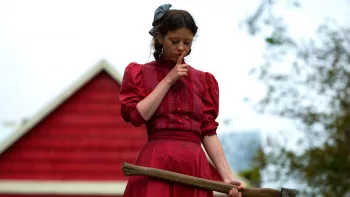 Mia Goth en robe rouge debout, le regard vers le sol, une pioche dans la main, un doigt sur les lèvres dans le film Pearl.