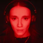 Une jeune femme, casque audio sur la tête, semble captivée, un peu inquiète, par quelque chose ; son visage est inondé dans une brutale lumière rouge alors que le fond est noir ; plan issu du film Les Chambres Rouges de Pascal Plante.