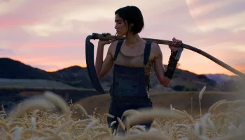 L'actrice Sofia Boutella dans un champ au crépuscule, tient une faucille sur ses épaules ; la tête baissée, elle a l'air mélancolique ; plan issu du film Rebel Moon - Partie 1 : Enfant du Feu.