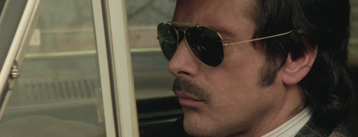 Tomas Millian l'air fermé, au volant de sa voiture à l'arrêt, portant des lunettes de soleil opaques ; plan du film Les féroces édité dans le coffret Les années de plomb volume 3.