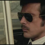 Tomas Millian l'air fermé, au volant de sa voiture à l'arrêt, portant des lunettes de soleil opaques ; plan du film Les féroces édité dans le coffret Les années de plomb volume 3.