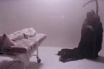 La Faucheuse s'approche d'un corps sous un drap dans une chambre mortuaire.