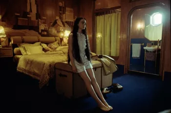 Emma Stone assise au pied de son lit, en chemise de nuit, pensive ; dans une chambre ancienne aux couleurs dorées présente dans le film Pauvres Créatures.