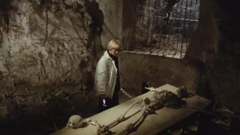 Une femme dans une crypte observe un squelette attaché à une planche en bois ; scène du film Le chat noir.