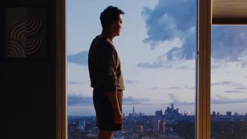 Andrew Scott est debout pensif devant une grande fenêtre qui donne sur une ville à building, ambiance de crépuscule ; plan issu du film Sans jamais nous connaître.