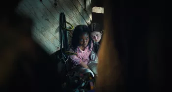 Deux petites filles angoissées sont assises côte à côte et regardent en direction de l'objectif, elles sont vues à travers une fente dans un mur ; plan du film L'Exorciste : Dévotion.