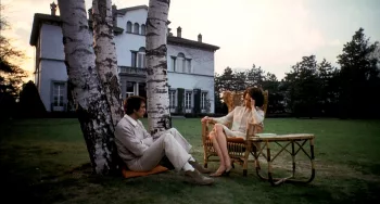 Terence Stamp assis au pied d'un arbre discute avec Silvana Mangano installée sur une chaise de jardin, sur l'herbe devant la grande maison du film Théorème.