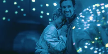 Deux amoureux rient en s’enlaçant, lors d'une soirée éclairée par des boules à facette, et plongée dans une lumière bleue claire ; ils semblent être les seules personnes de l'endroit.