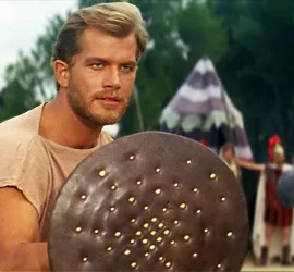 Richard Harrison dans Persée L'invincible prêt à un combat d'entraînement, derrière un bouclier et une rangée de soldats qui observent.