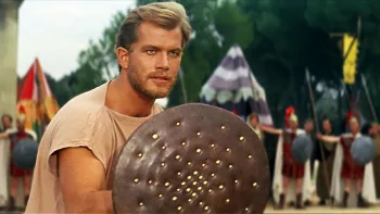 Richard Harrison dans Persée L'invincible prêt à un combat d'entraînement, derrière un bouclier et une rangée de soldats qui observent.