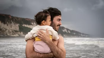 Payanotis Pascot torse nu, tient son bébé dans les bras, tous deux ont le visage tourné vers la mer et l'horizon, sous un ciel gris et devant les falaises ; scène de la série De Grâce.