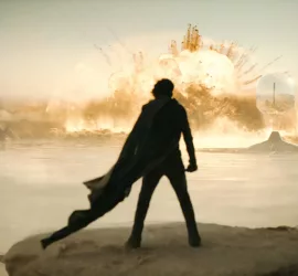 Vue de dos, la silhouette de Timothée Chalamet portant une longue cape noire, observe une large explosion dans ce qui semble être un désert ; plan issu du film Dune : deuxième partie.