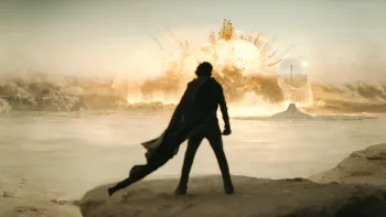 Vue de dos, la silhouette de Timothée Chalamet portant une longue cape noire, observe une large explosion dans ce qui semble être un désert ; plan issu du film Dune : deuxième partie.