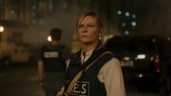 Plan rapproché-taille sur Kristen Dunst, vête d’une veste indiquant qu'elle fait partie du service de presse, qui observe dans une rue de nuit, au loin, soucieuse, dans le film Civil War.