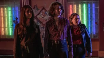Trois adolescentes personnages du film Drive-Away Dolls semblent inquiètes, dans un lieu indéfini, aux murs beiges et aux fenêtres condamnées avec des néons de toutes les couleurs ; toutes les trois côte à côte, regardant vers leur gauche, dans l'appréhension de ce qui pourrait venir.