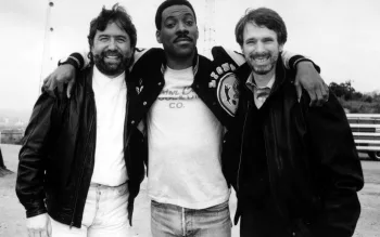 Eddie Murphy pose bras dessus bras dessous avec Jerry Bruckheimer (à sa gauche) et Don Simpson (à sa droite) sur le tournage du Flic de Beverly Hills, tous les trois tout sourire photographie en noir et blanc.