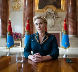 Kate Winslet en présidente, assise dans une salle luxueuse, les mains jointes, attentive ; derrière elle, cinq drapeaux du pays fictif de la série The Regime.
