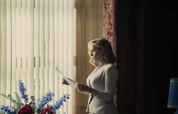 Kate Winslet, debout près d'une large fenêtre lumineuse à peine voilée par des stores, lit une lettre, vêtue d'une robe blanche sobre et élégante, dans la série The Regime.