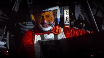 Dave Filoni en pilote spatial rebelle, dans le cockpit dans son vaisseau, en casque et uniforme orange et blanc.