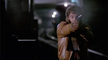 Plan rapproché-taille sur un homme en veste de cuir marron qui tire avec un revolver, sur les rames du métro déserts ; scène du film Les rats attaquent.