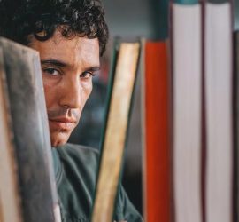 Gros plan sur le visage du comédien Adam Bessa, méfaitn, observant entre les livres d'un rayon de bibliothèque dans le film Les fantômes.