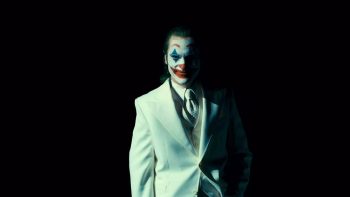 Le Joker, en costume cravate blancs se tient droit, les mains dans les poches, dans la pénombre, seul lui est éclairé ; plan du film Joker : folie à deux.