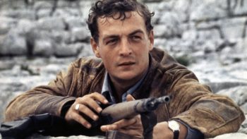 Philippe Léotard regarde l'horizon, un fusil entre les mains, devant une paroi de pierres grises dans le film Le franc-tireur.