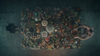 Plan en plongée zénithale sur une grande table remplie de mets et de nourriture ; un homme et une femme sont d'un bout à l'autre ; issu du film La plateforme 2.