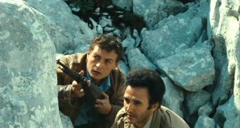 Deux hommes sont cachés derrière des roches grises, armés, dans le film Le franc-tireur.