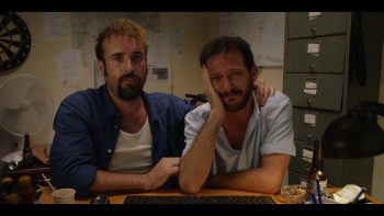 Deux hommes dans une salle d'interrogatoire, face caméra ; tous deux semblent venir d'une classe sociale défavorisée, à la campagne ; l'un des deux hommes a son bras autour des épaules de l'autre à côté de lui, en train de pleurer : plan issu du film Les pistolets en plastique.