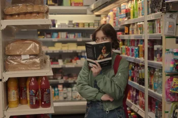 Une jeune femme se avec avec un livre sur le visage dans les rayons d'une pharmacie ; scène de Meurtre mode d'emploi.