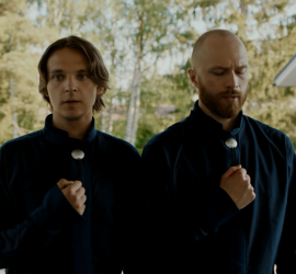 Deux hommes, sur le palier d'une maison en résidence pavillonnaire, vêtues d'une chemise noire, semblent prier, la main sur la poitrine, dans le film The Redeemers.