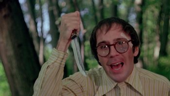 Un jeune homme au look d'étudiant introverti (lunettes imposantes, chemise fermée jusqu'au col) brandit un couteau en pleine forêt ; scène du film I spit on your grave.